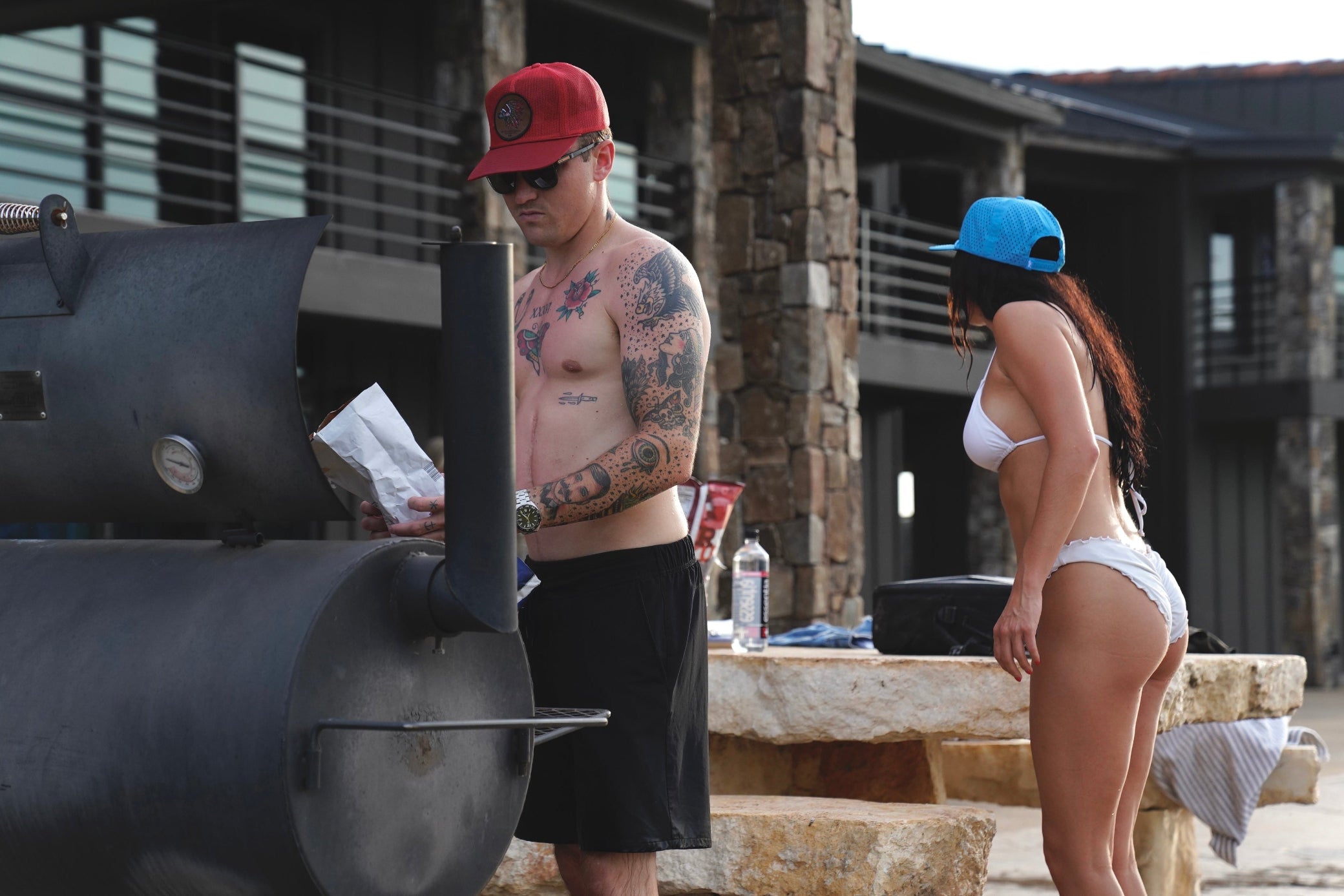 Man with tattoos and a woman in a bikini standing near a barbecue grill outdoors.